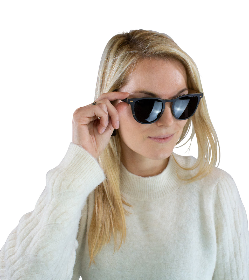 Closeup of female model in wood wayfarer sunglasses, adjusting arm with one hand: gold corner accents, dark non-mirror lenses