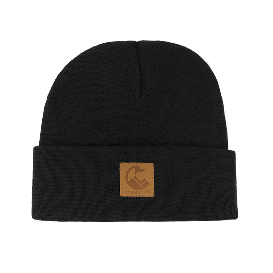 Front view of black, acrylic knit beanie: wide rollover cuff with Conquer Life logo patch in caramel faux-leather