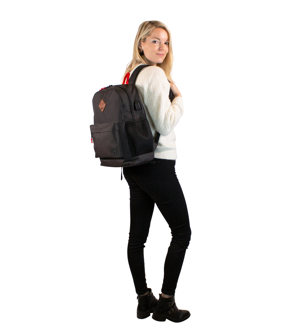 Rear view of standing female model wearing black poly-fabric backpack strapped over 1 shoulder, turning face to camera