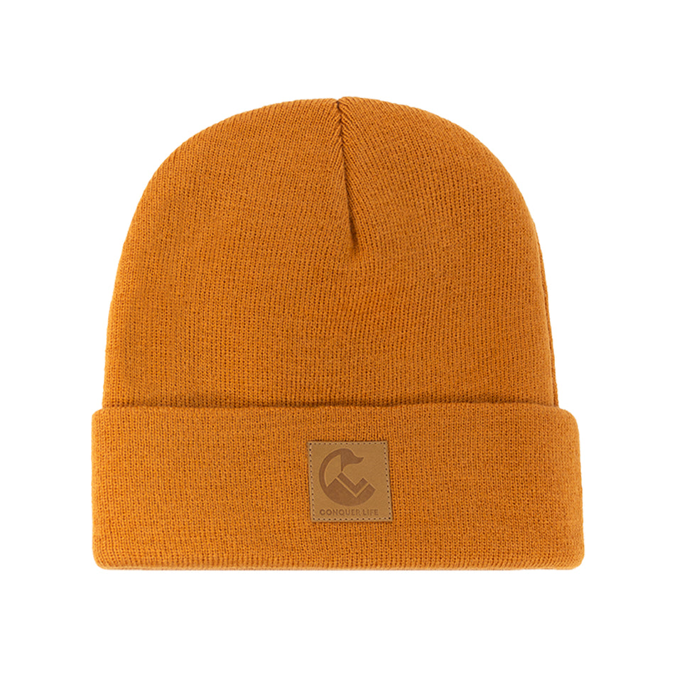 Front view of brown, acrylic knit beanie: wide rollover cuff with Conquer Life logo patch in caramel faux-leather