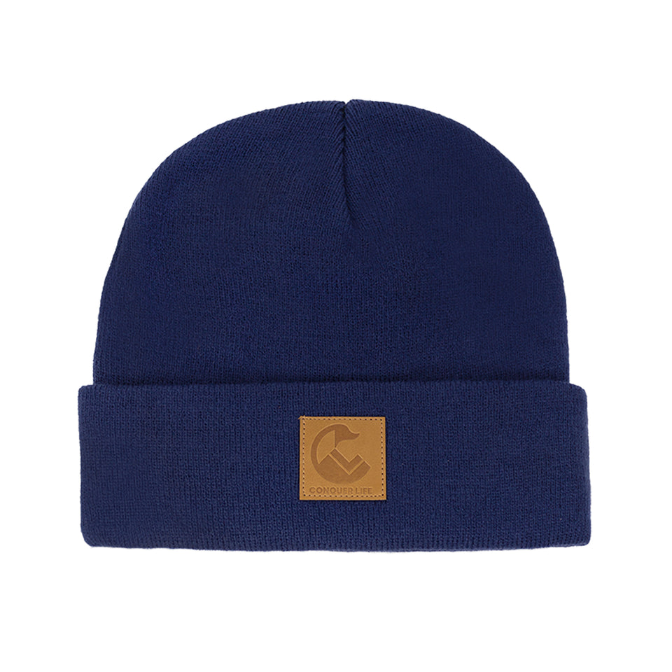 Front view of navy blue, acrylic knit beanie: wide rollover cuff with Conquer Life logo patch in caramel faux-leather