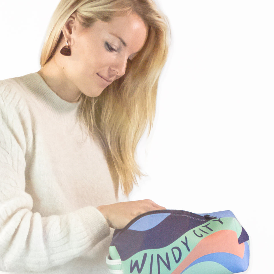Female model from waist-up, her hand in unzipped neoprene pouch, showing long side with multicolor waves & Windy City text