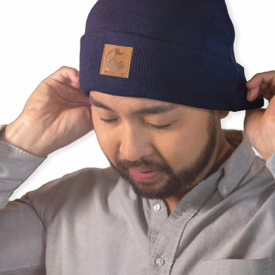 3/4 front, shoulders-up view of male model in navy blue beanie: his hands folding the cuff, faux-leather patch is off-center 