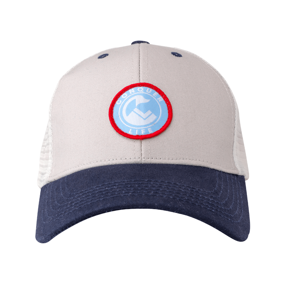 Front of light gray trucker hat with blue bill: circular, red-rimmed light blue patch with white Conquer Life text & logo 